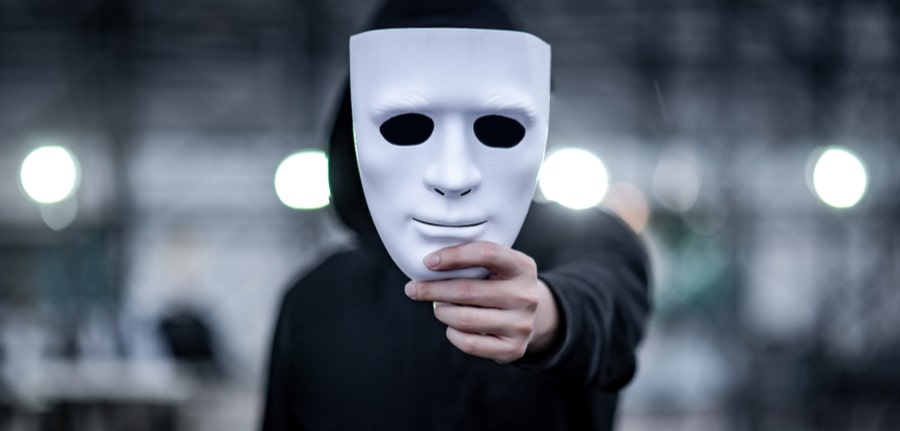 Mystery hoodie man with black mask holding white mask in his hand depciting a hacker during Halloween Cybersecurity concept