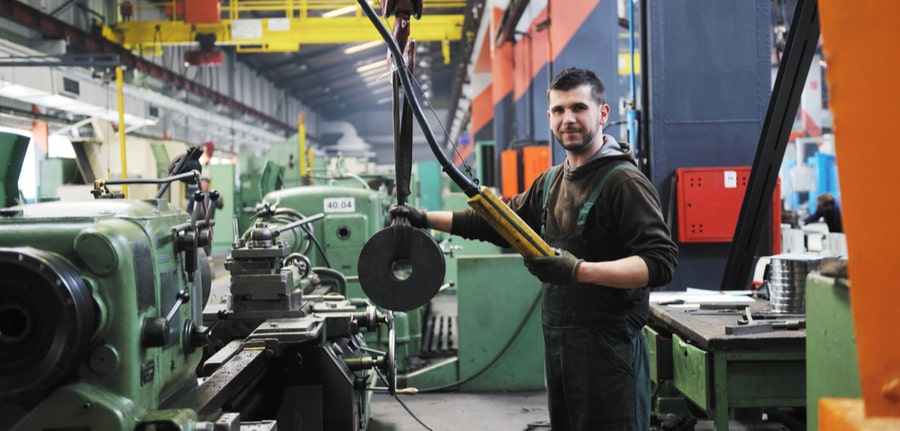 A factory worker in a manufacturing plant full of machinery, depicinting computer technology and cybersecurity in industrial firms